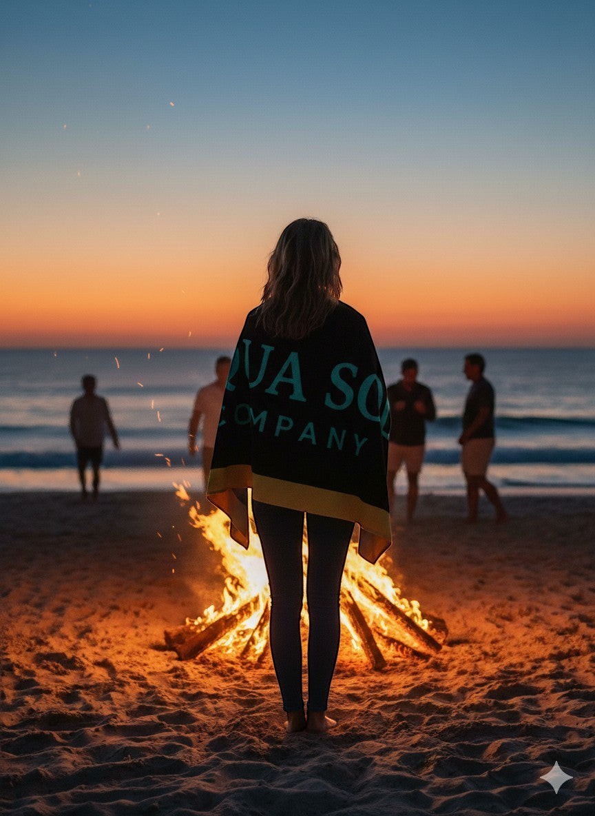 Aqua Sol Company beach towel draped over a woman standing before a beach bonfire at sunset with friends nearby