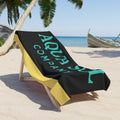 Aqua Sol Company beach towel in black and yellow with aqua branding, draped over a wooden lounger on a sandy beach.