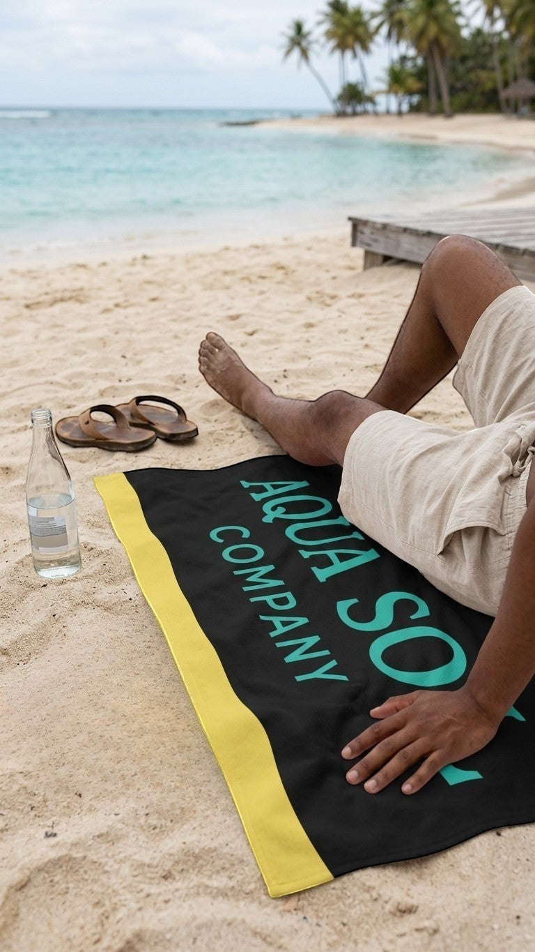 Aqua Sol Company beach towel with black, yellow, and aqua branding, laid on sand with a person relaxing nearby.