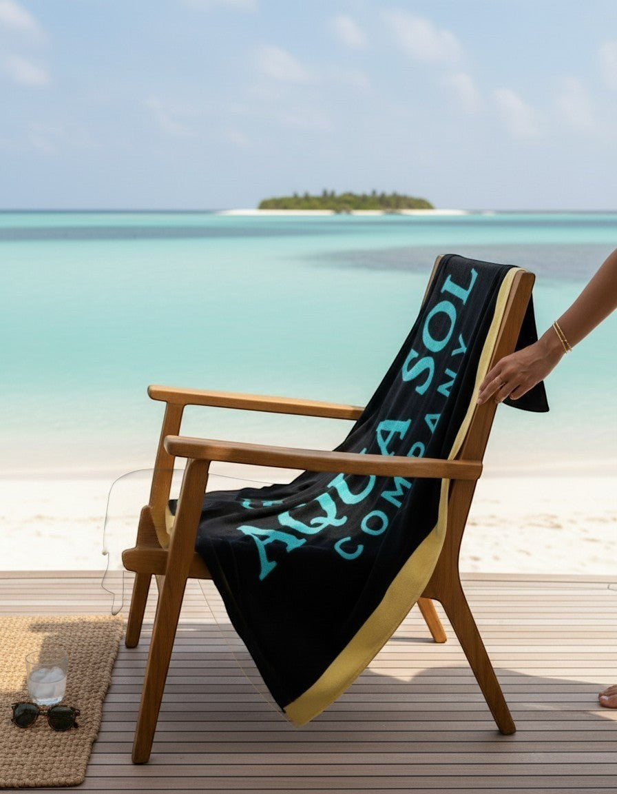 Aqua Sol Company beach towel in black with aqua logo draped over a wooden chair on a tropical beach deck